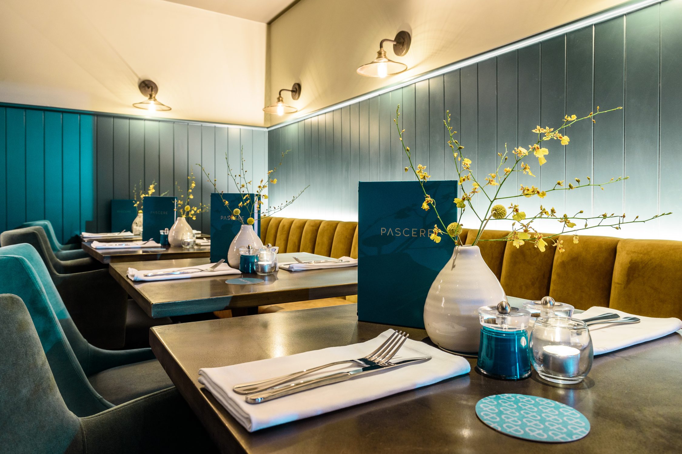 Restaurant interior design including wall lights illuminating a wall, half clad with blue v-groove panelling. In the foreground dark wood tables are dressed ready for dinner. Stickland Wright Architects and Interior Design, Brighton & Hove