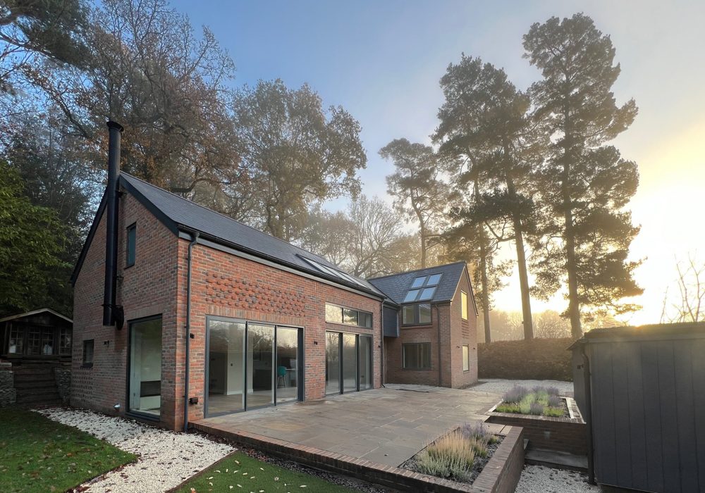 Low Energy Home Visualisation with brick walls, large stretches of glazing and zinc roof. The house is surrounded by tall trees and a patio area stretches out in front - Stickland Wright Architects Brighton