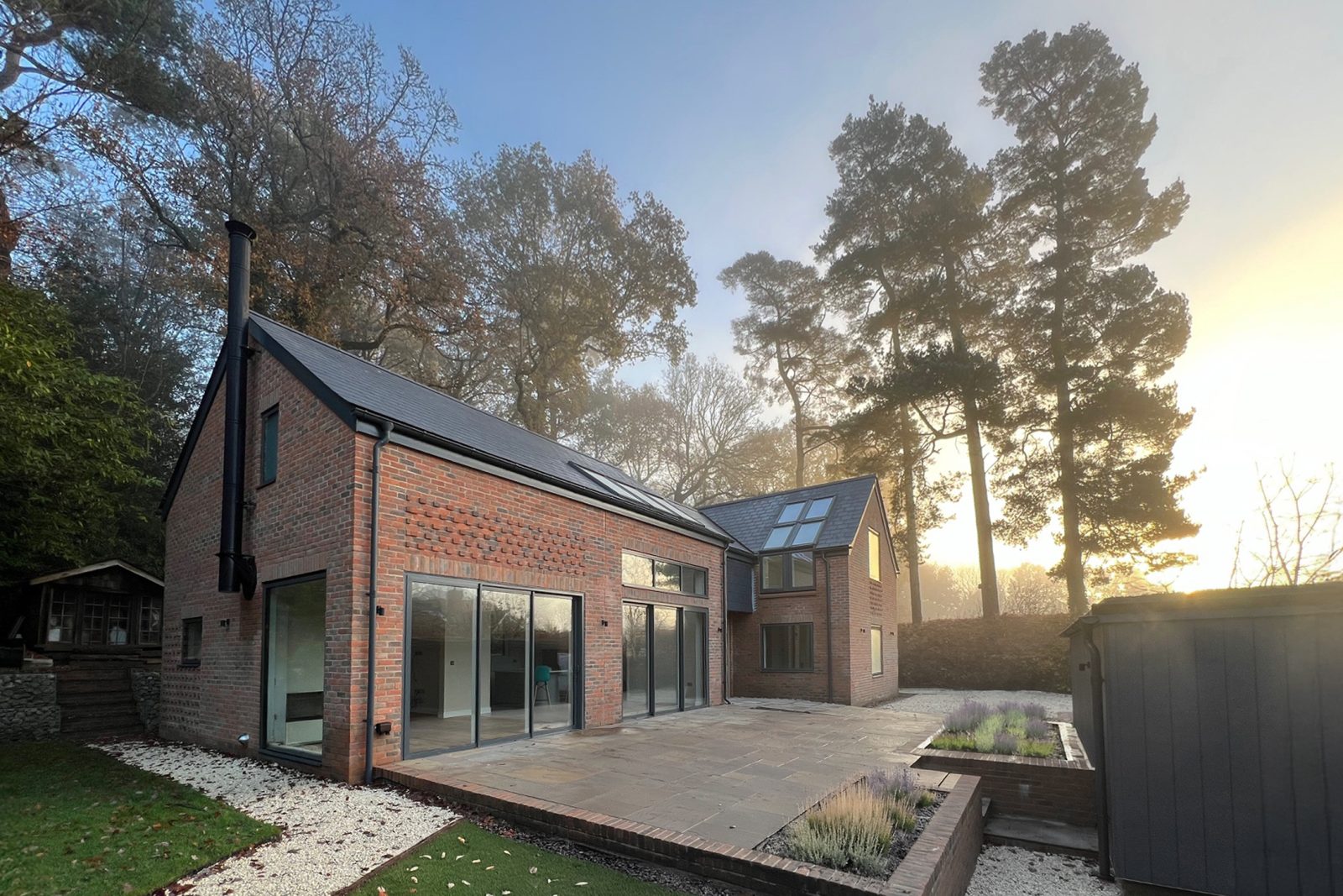 Low Energy Home Visualisation with brick walls, large stretches of glazing and zinc roof. The house is surrounded by tall trees and a patio area stretches out in front - Stickland Wright Architects Brighton