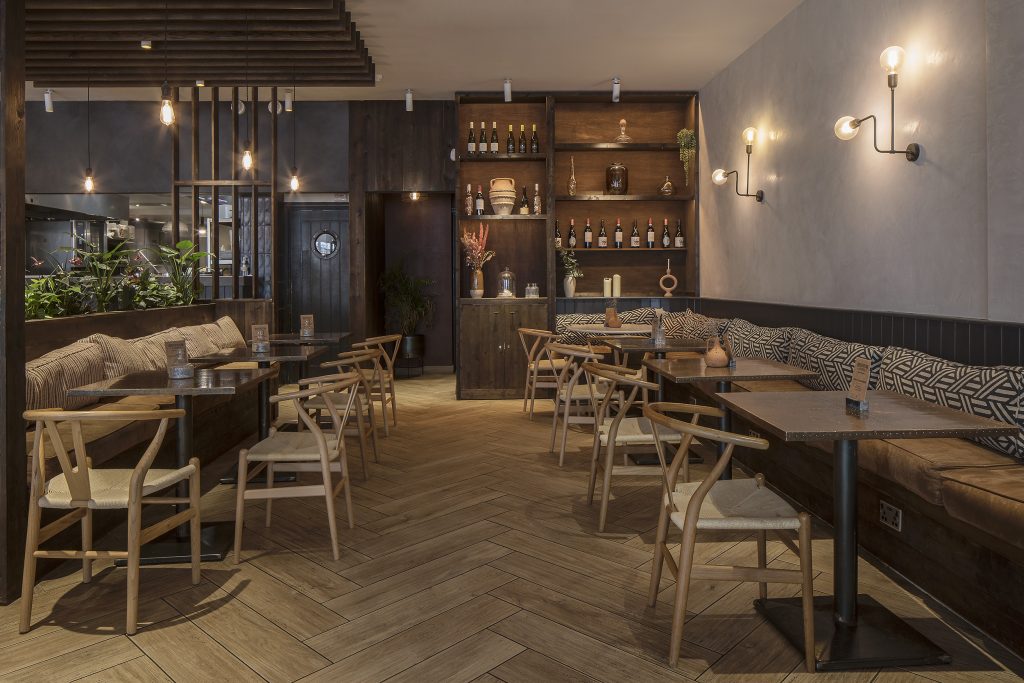 View of a café interior including relaxed banquette seating to the sides. Wishbone style loose chairs face the banquet. Bespoke dark wood joinery includes shelving at the rear and a partition to the left. Stickland Wright Architects and Interior Design, Brighton & Hove