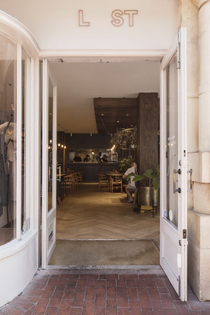 View through open double doors into a cafe interior. The design includes herringbone flooring and dark wood wall cladding. Stickland Wright Architects and Interior Design, Brighton & Hove